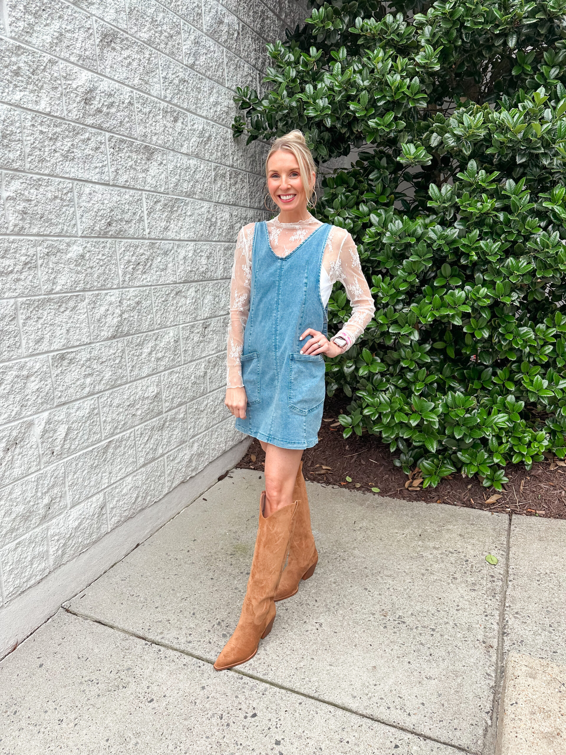 Woman smiling in a casual-chic outfit featuring a sleeveless denim dress layered over a sheer white lace shirt, styled with tan suede cowboy boots. She’s standing on a sidewalk beside a white brick wall and lush green bushes.